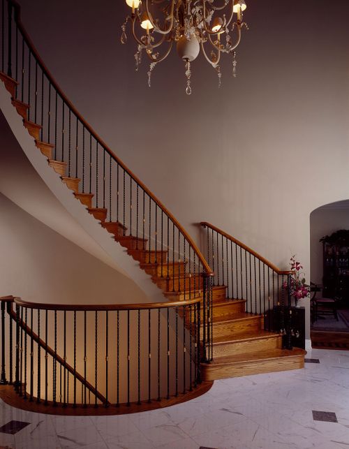 Elegant wooden staircase with black wrought iron railing, illuminated by a chandelier, set in a spacious foyer.