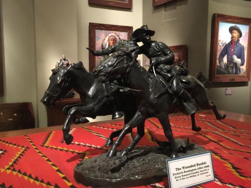 A bronze sculpture titled "The Wounded Bunkie," depicting two cowboys on horseback, set against a colorful backdrop.