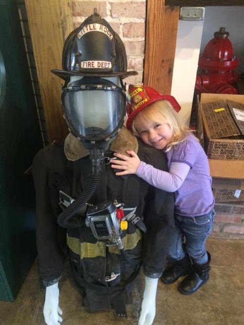 A young girl in a red fire hat hugs a firefighter mannequin in a cozy setting.