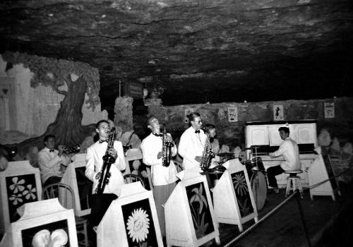 A vintage band performs in a cave-like venue, featuring musicians with saxophones and a pianist at a white piano.