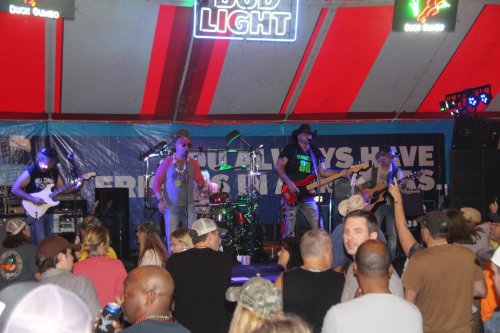 A lively band performs on stage under a colorful tent, with an engaged crowd enjoying the music.