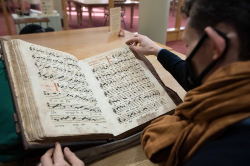 A person examines an open, antique music book with handwritten sheet music in a library setting.