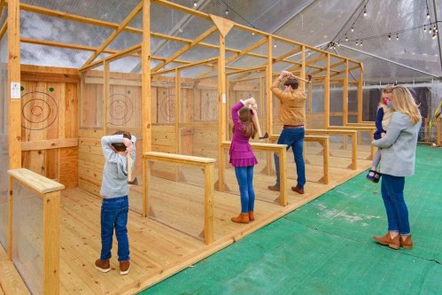 A family stands in an indoor axe-throwing range, preparing to throw axes at wooden targets.