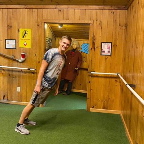 A young man in a t-shirt and shorts leans playfully in a hallway, with a mannequin in a coat standing in the background.