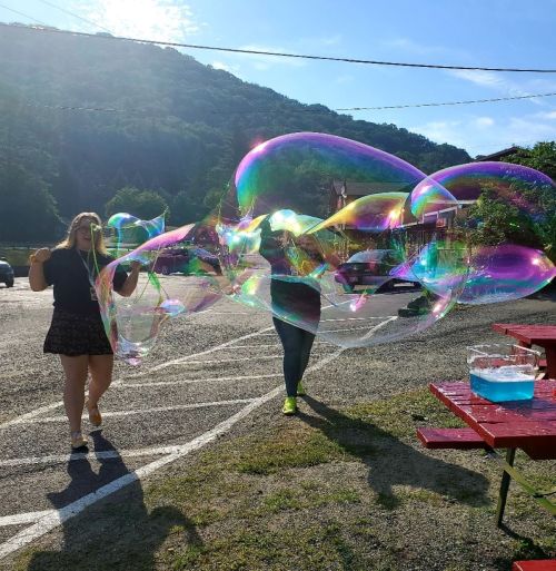 Two people create large, colorful soap bubbles in a sunny outdoor setting, with trees and a blue sky in the background.