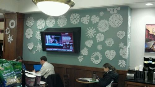 A cafe interior with a wall decorated in paper snowflakes and a TV displaying news, while patrons work on laptops.