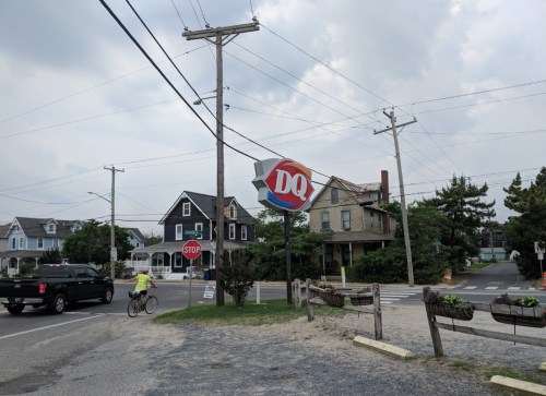 A Dairy Queen sign near a stop sign, with a cyclist and houses in the background under a cloudy sky.