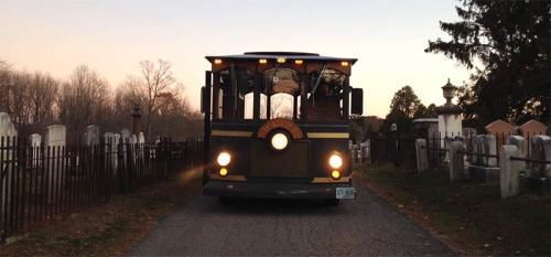 A vintage trolley approaches along a path lined with gravestones and trees at dusk.