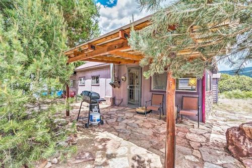 A cozy outdoor patio with a grill, surrounded by trees and stone flooring, leading to a small house.