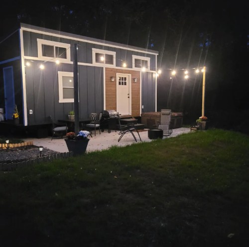 A cozy cabin at night, illuminated by string lights, with a patio area and potted plants.