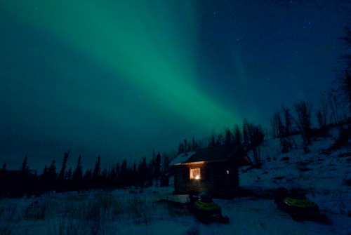 A cozy cabin illuminated by a warm light under a vibrant green aurora borealis in a snowy landscape.