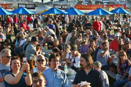 A large crowd enjoying an outdoor festival with blue umbrellas and food stalls in the background.