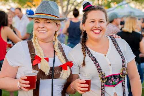 Two women in traditional attire hold drinks at an outdoor festival, smiling and enjoying the event.