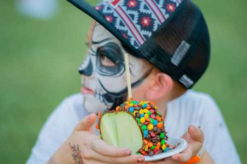 A child with face paint holding a caramel apple covered in colorful candies, looking off to the side.