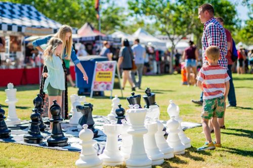 Children play with large chess pieces at an outdoor market, surrounded by colorful tents and a lively atmosphere.