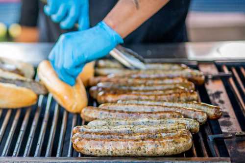 A person wearing blue gloves is grilling sausages and preparing hot dog buns on a barbecue grill.