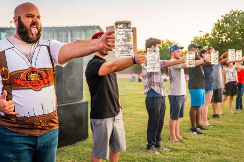 A group of people in a park holding up beer steins, with one man in a costume playfully sticking out his tongue.