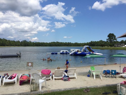A sandy beach by a lake with inflatable water toys, people swimming, and lounge chairs under umbrellas.