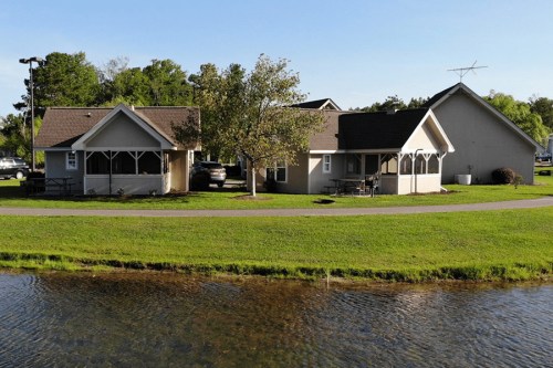Two charming houses by a calm lake, surrounded by green grass and trees under a clear blue sky.
