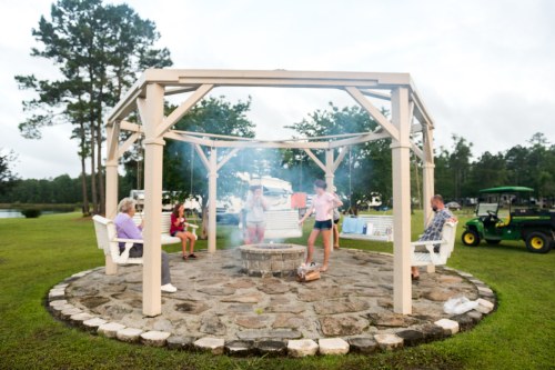 A group of people gather around a stone fire pit under a gazebo, with trees and a tractor in the background.