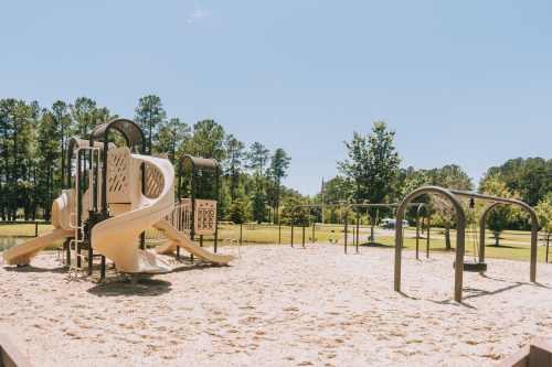 A playground with a slide and swings, surrounded by trees and a clear blue sky. Sand covers the ground.