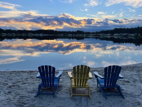 Three colorful adirondack chairs on a sandy beach, facing a calm lake reflecting a vibrant sunset sky.