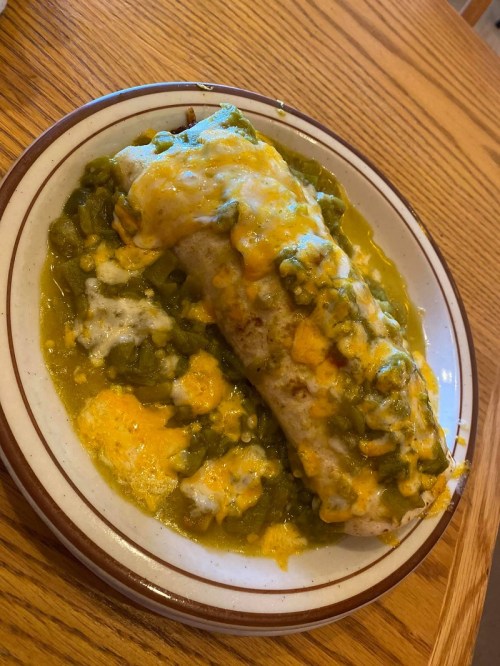 A plate of a cheesy burrito topped with green sauce and melted cheese, served on a wooden table.