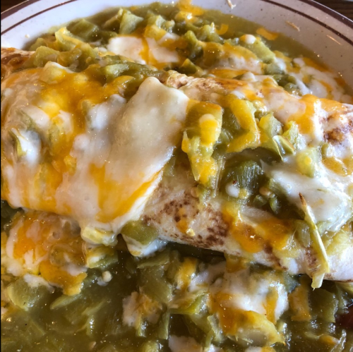 A close-up of a cheesy enchilada topped with green sauce and diced green chilies on a plate.
