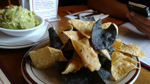 A plate of mixed tortilla chips, both yellow and blue, with a bowl of guacamole in the background.