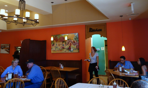 A cozy restaurant interior with wooden tables, colorful artwork, and a server walking towards a dining area.