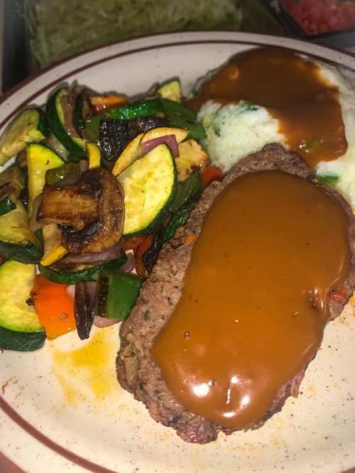 A plate featuring meatloaf with gravy, mashed potatoes, and a side of sautéed vegetables including zucchini and bell peppers.