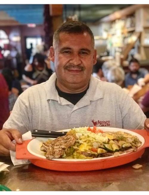 A smiling man holds a large platter of food, featuring grilled meat and colorful vegetables, in a busy restaurant setting.