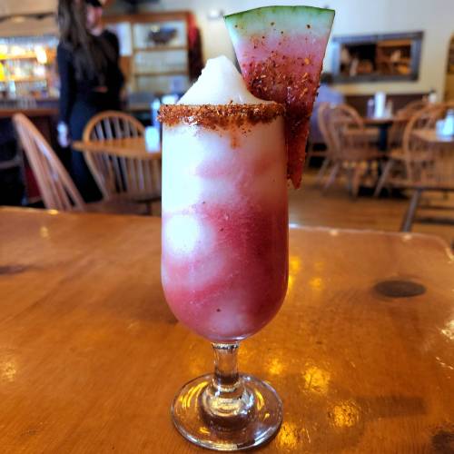 A colorful frozen drink in a tall glass, garnished with a watermelon slice and chili powder on the rim.