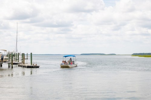 A boat with passengers navigates a calm waterway near a dock under a cloudy sky.