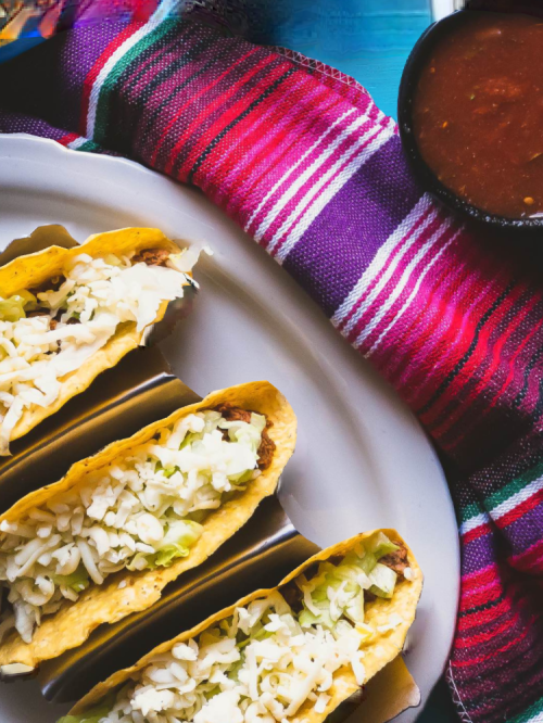 Three tacos filled with lettuce and cheese on a plate, accompanied by a bowl of salsa and a colorful woven blanket.