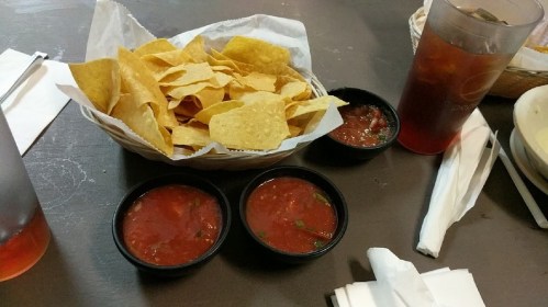 A basket of tortilla chips with two bowls of salsa and a glass of iced tea on a table.