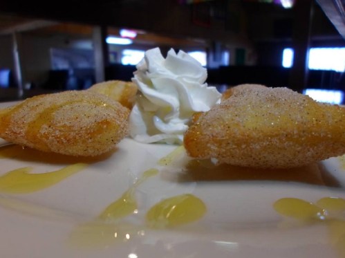 A close-up of two golden pastries dusted with sugar, served with whipped cream and a drizzle of sauce on a plate.