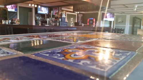 A close-up of a colorful tiled bar top with a blurred background of a bar and two TVs.