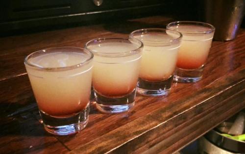 Four shot glasses lined up on a wooden surface, filled with a layered drink featuring yellow and reddish hues.