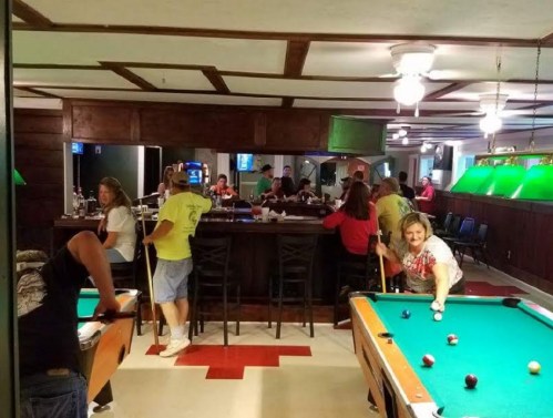 A lively bar scene with people playing pool and socializing, featuring green pool tables and a wooden interior.