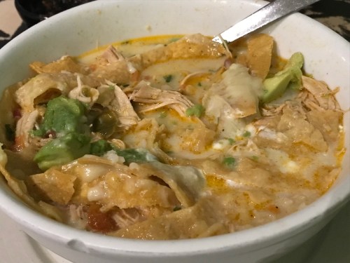 A bowl of chicken tortilla soup with shredded chicken, tortilla strips, avocado, and melted cheese.