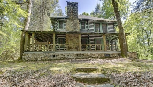 A rustic log cabin surrounded by trees, featuring a stone chimney and a spacious porch.
