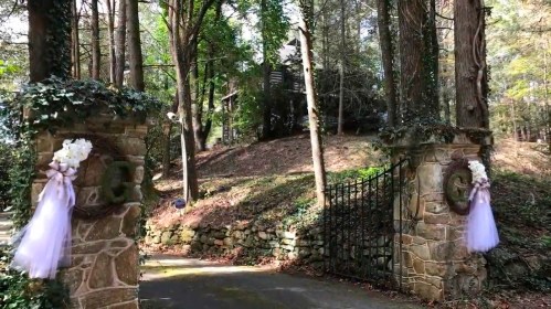 A scenic entrance with stone pillars adorned with white flowers and tulle, leading to a wooded path.