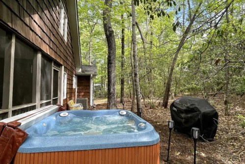 A hot tub sits beside a house, surrounded by trees and a grill, creating a serene outdoor retreat.