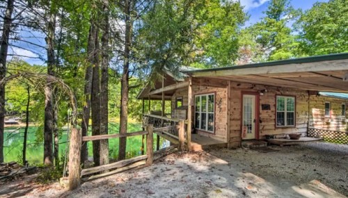 A rustic cabin surrounded by trees, with a view of a green lake and a wooden deck.
