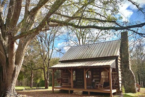 A rustic log cabin with a metal roof, surrounded by trees and a clear blue sky. Rocking chairs on the porch.