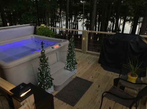 A cozy deck with a hot tub, two lit Christmas trees, and a grill, surrounded by trees and a serene lake view.