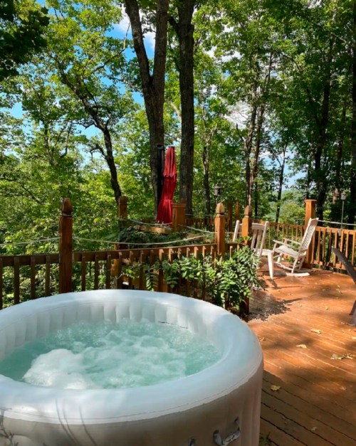 A hot tub on a wooden deck surrounded by lush green trees and a clear blue sky.
