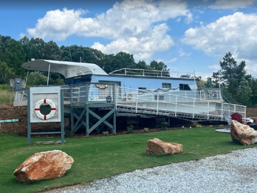 A large houseboat on a grassy area, with a life preserver nearby and rocky landscaping under a cloudy sky.