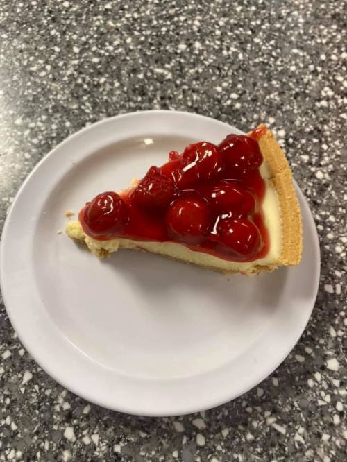 A slice of cherry cheesecake on a white plate, topped with bright red cherries and a graham cracker crust.